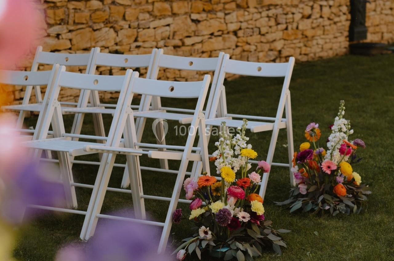 Chaises blanches pliantes alignées sur une pelouse, décorées de bouquets colorés devant un mur en pierre.