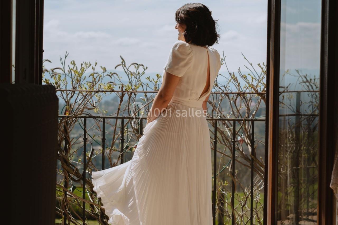Femme en robe blanche debout sur un balcon, regardant un paysage ensoleillé avec des collines verdoyantes.