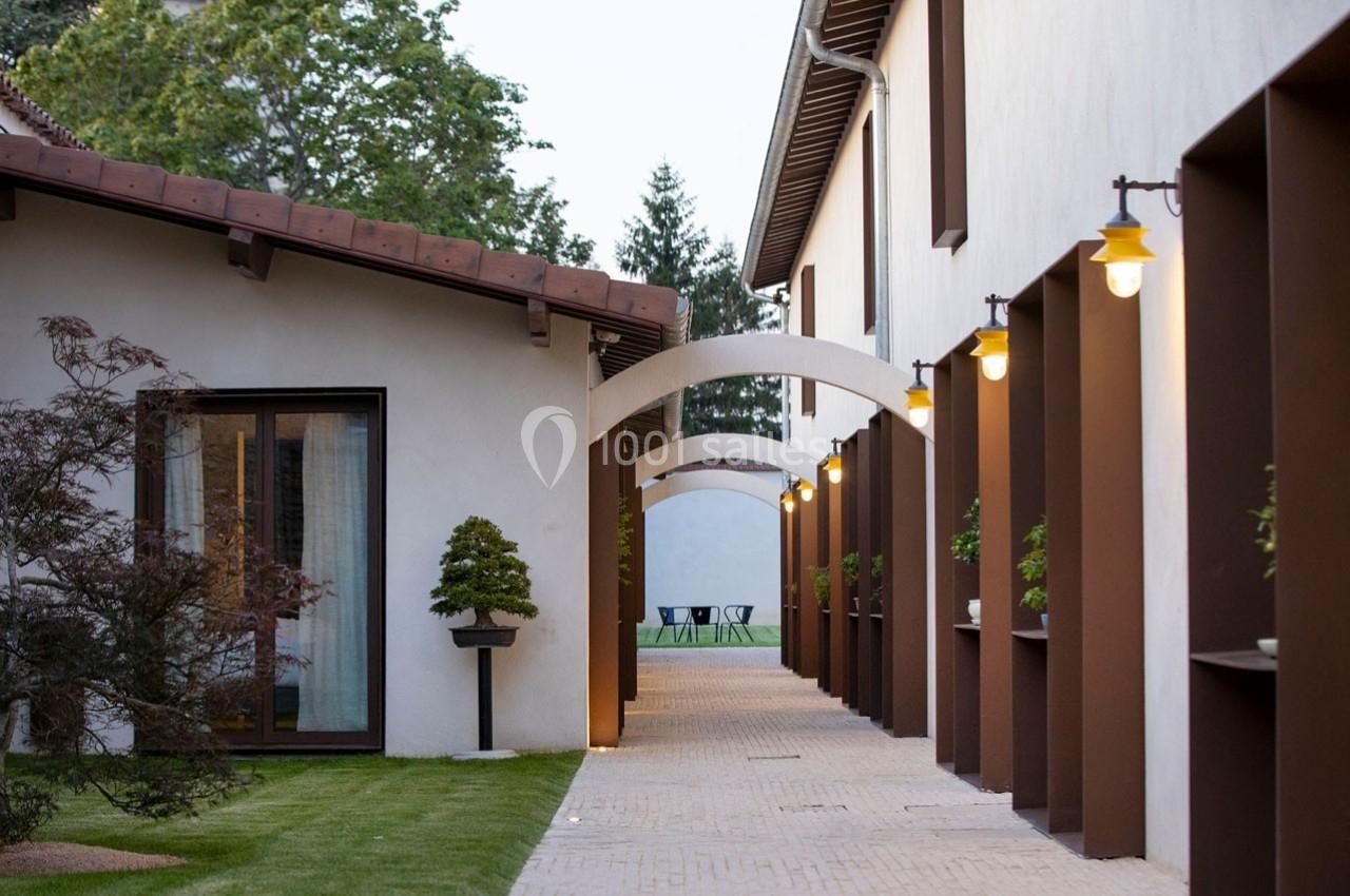 Passage extérieur bordé d'arches, éclairé par des lampes murales, avec vue sur un jardin et des chaises au fond.