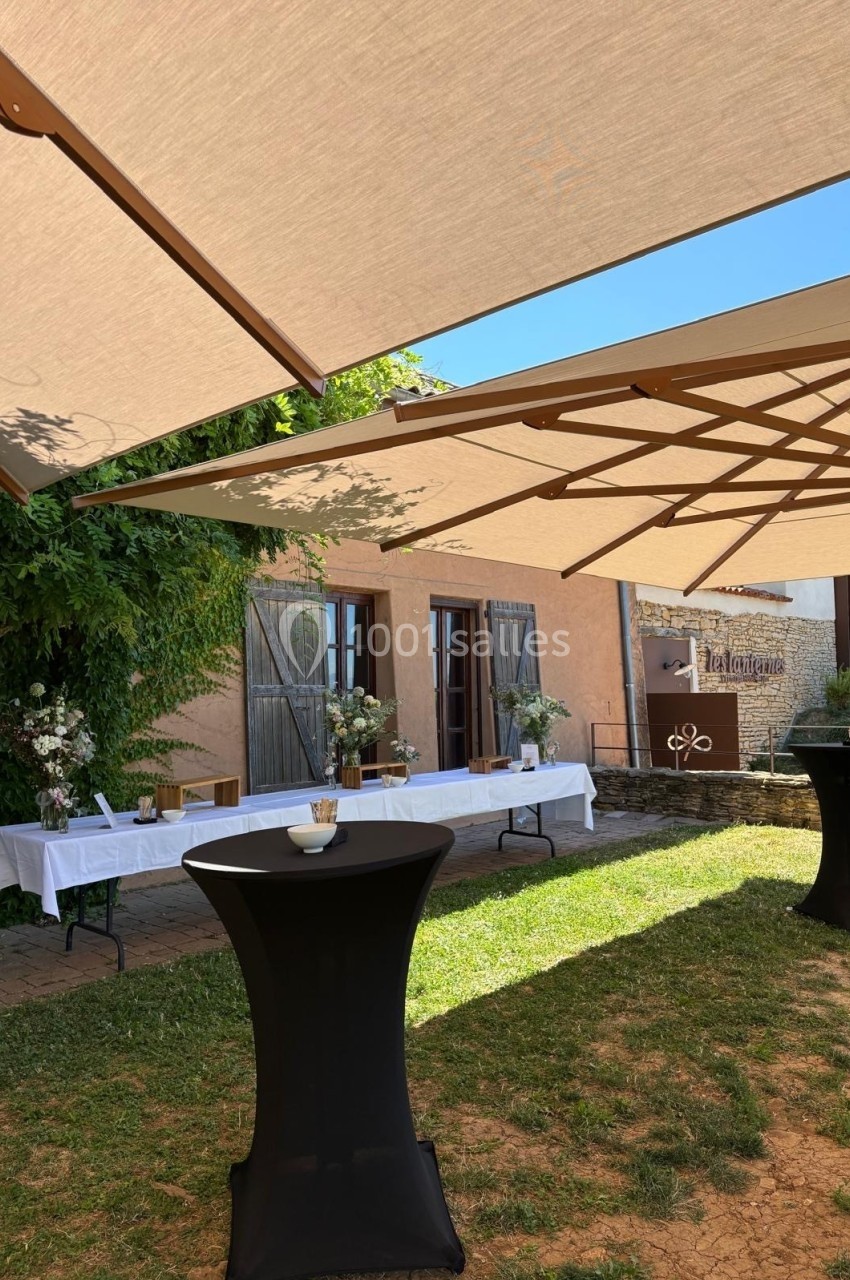 Terrasse ombragée avec des tables hautes et des buffets décorés, devant un bâtiment aux volets en bois.