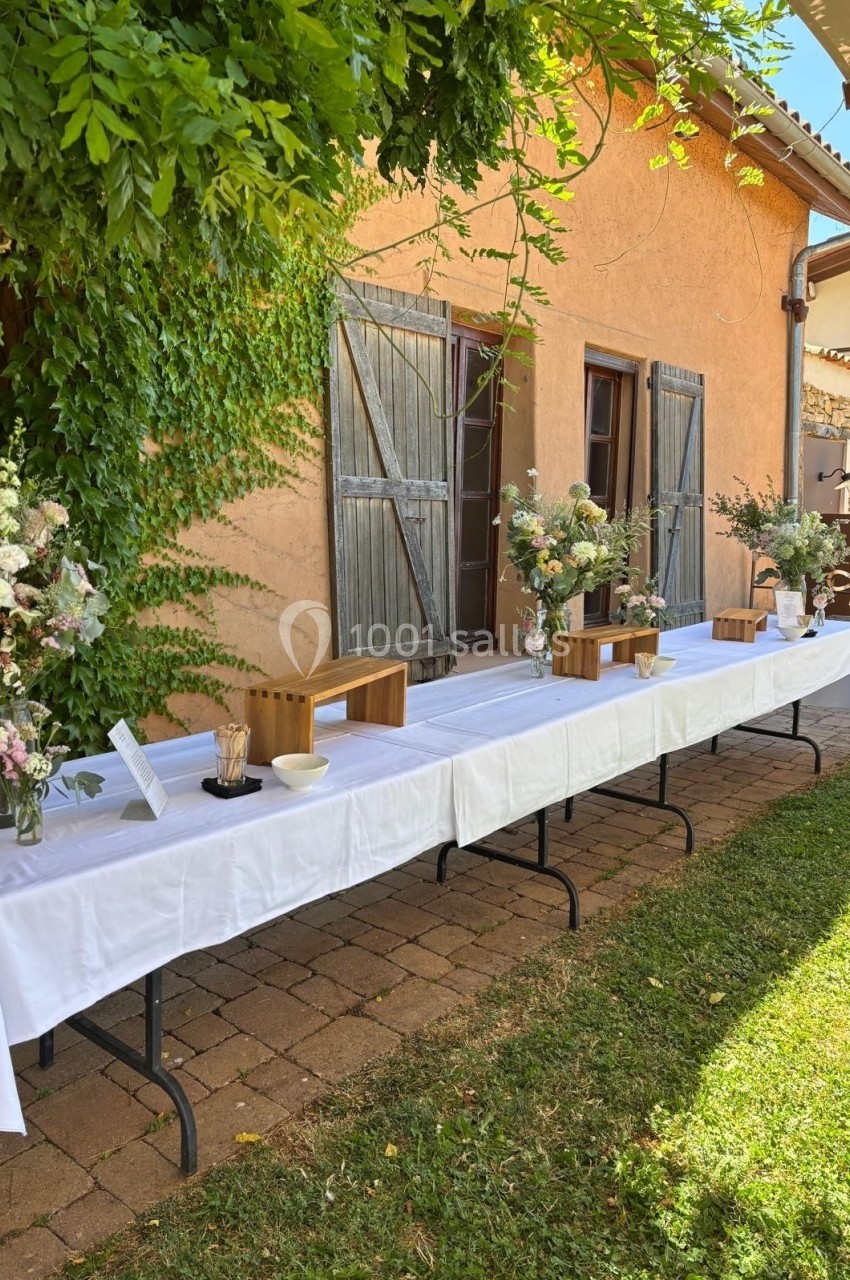 Table rectangulaire dressée en extérieur avec nappes blanches, décorée de fleurs et de boîtes en bois, près d'un mur…