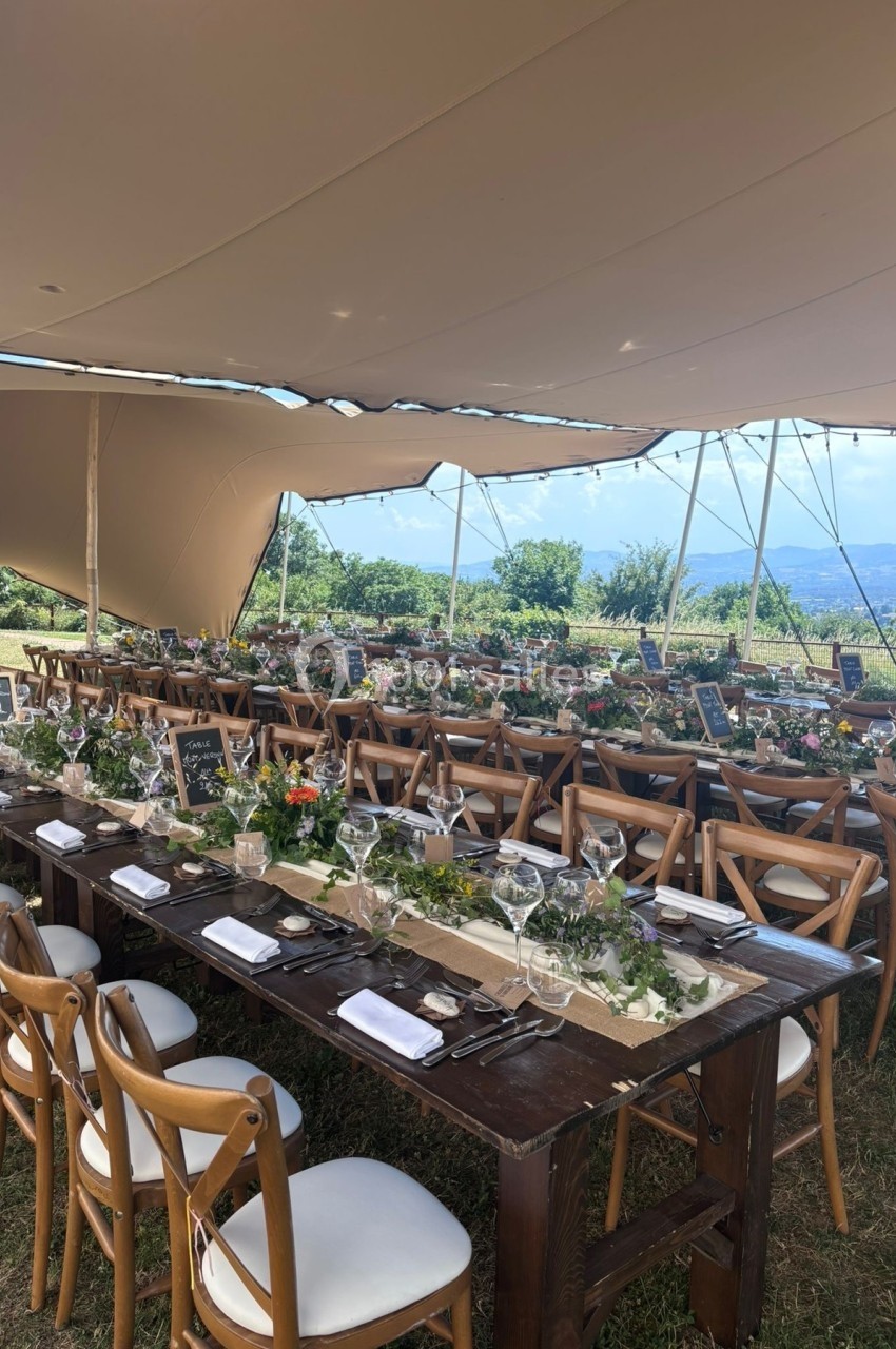 Tables en bois décorées de fleurs et de vaisselle sous une tente ouverte dans un cadre champêtre.
