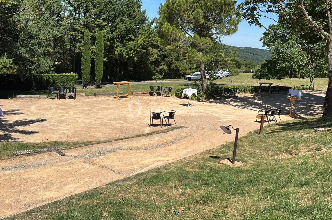 Espace extérieur avec tables et chaises disposées sur une place en terre, entourée de verdure et d'arbres.