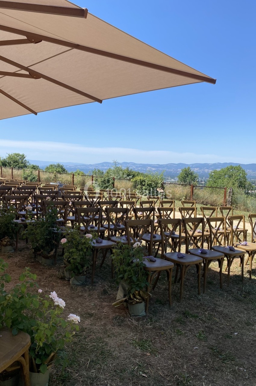 Chaises en bois alignées pour une cérémonie en extérieur sous un grand parasol, avec vue sur des collines verdoyantes.