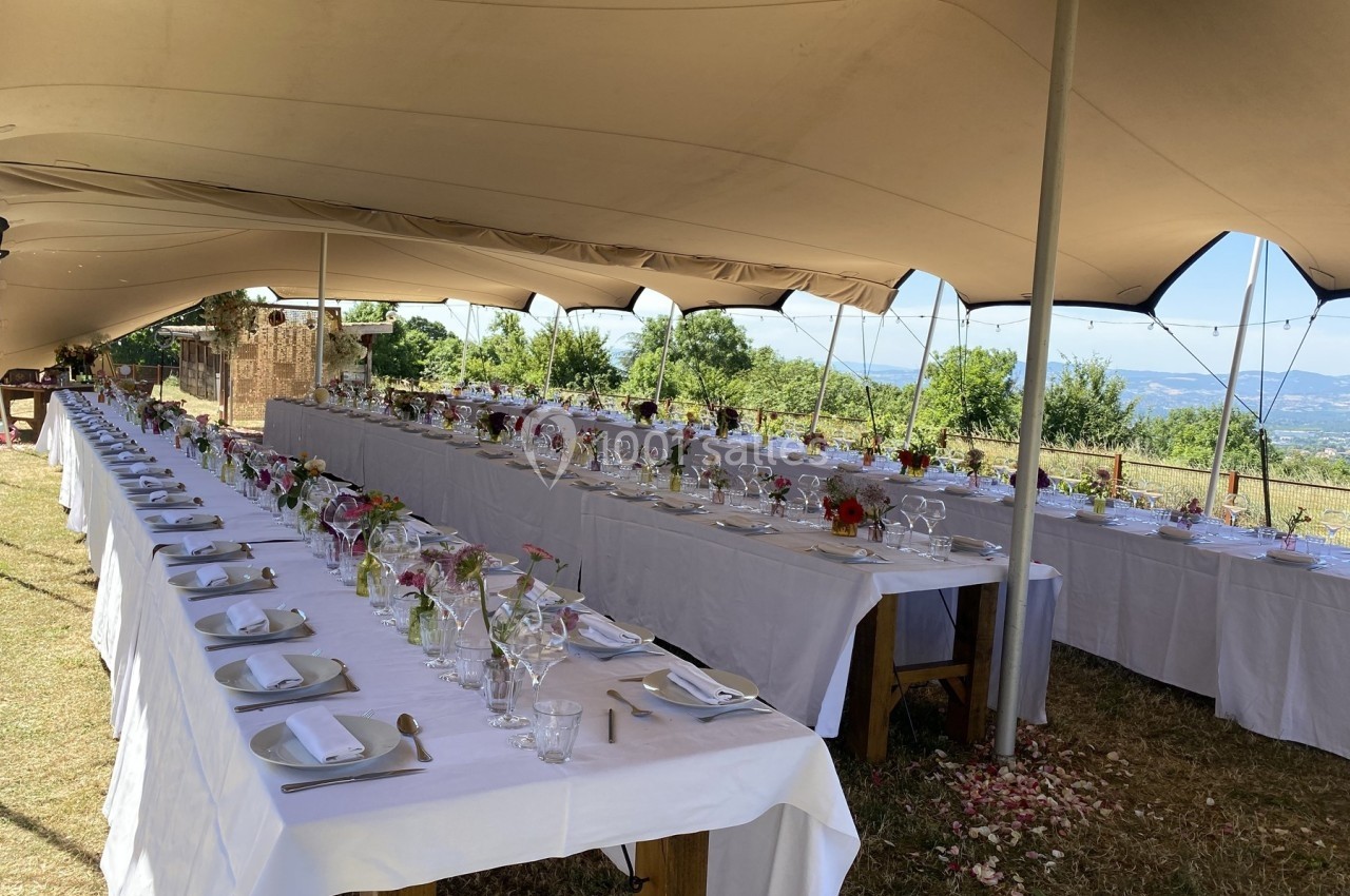 Tables longues dressées sous une tente, décorées de fleurs et de vaisselle, dans un cadre extérieur ensoleillé.