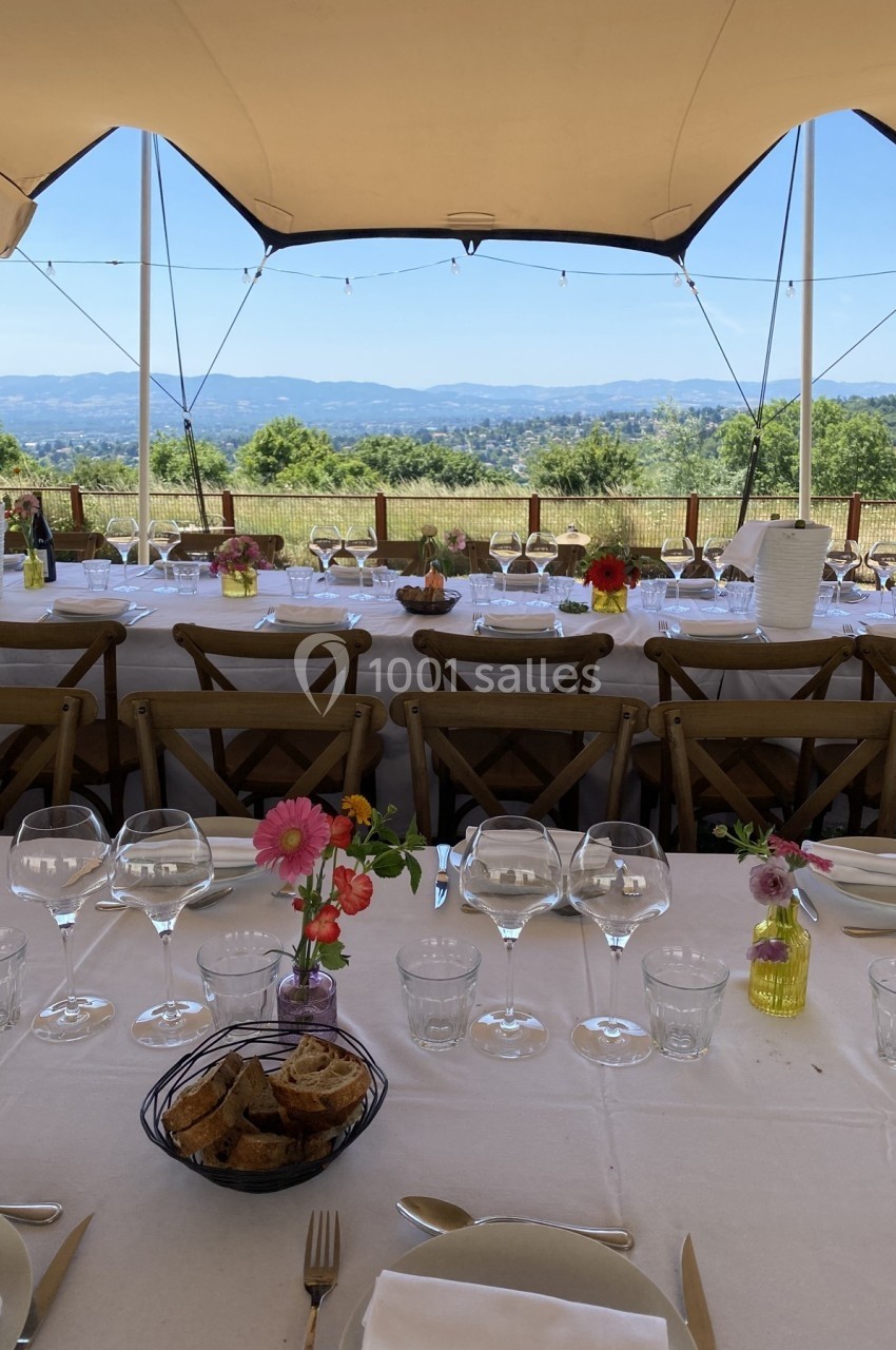 Tables dressées en extérieur sous une tente, avec vue sur un paysage vallonné et des décorations florales colorées.