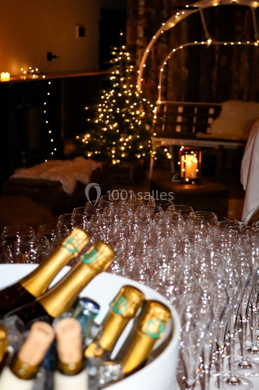 Bouteilles de champagne et verres alignés au premier plan, décor festif avec sapin illuminé en arrière-plan.