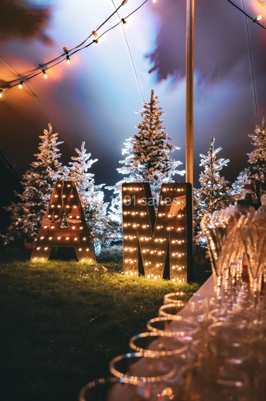 Lettres lumineuses ’AM’ devant des sapins décorés, sous une tente éclairée, avec des verres alignés au premier plan.