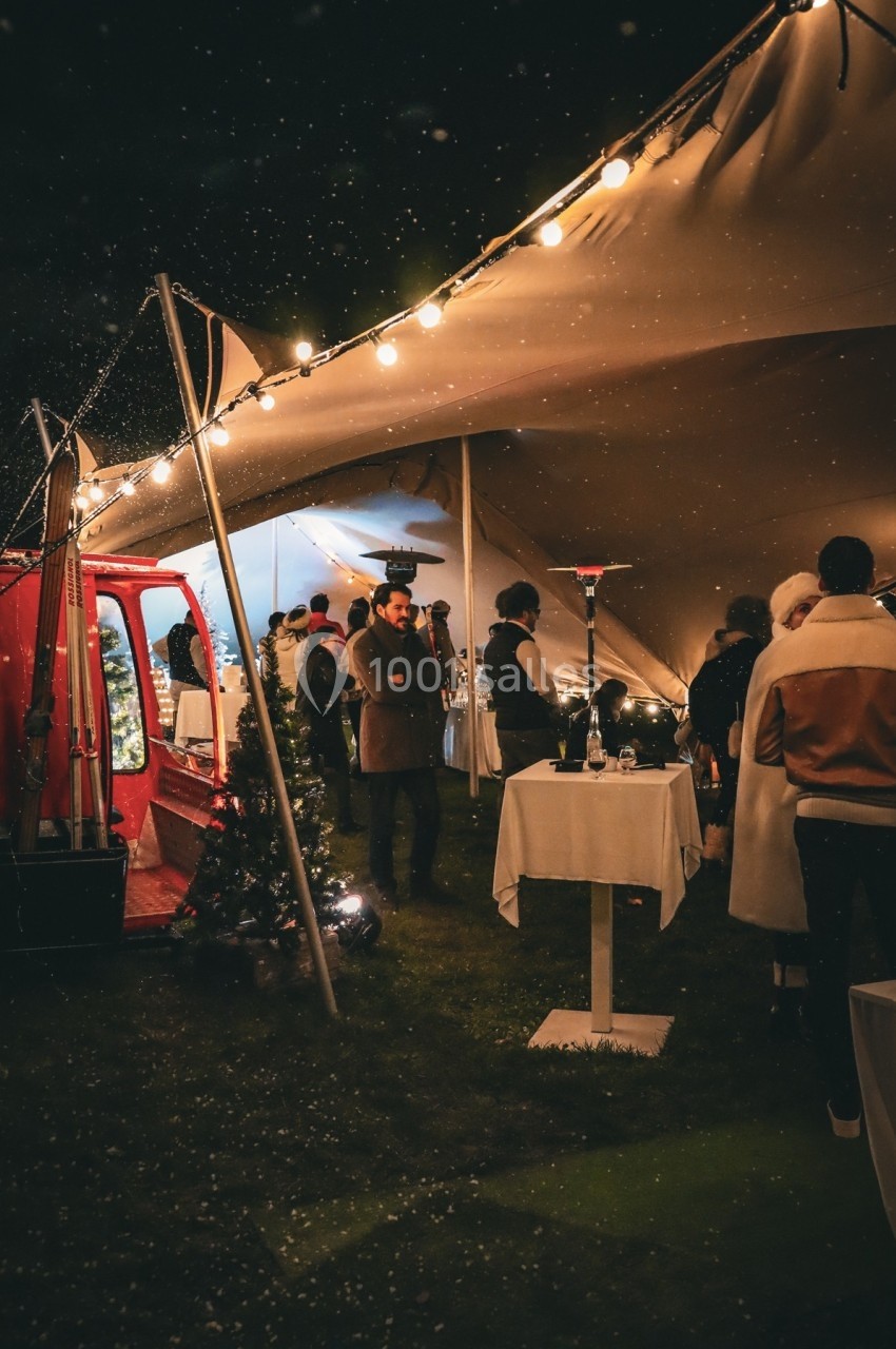 Personnes rassemblées sous une tente éclairée la nuit, avec tables hautes et décorations festives.