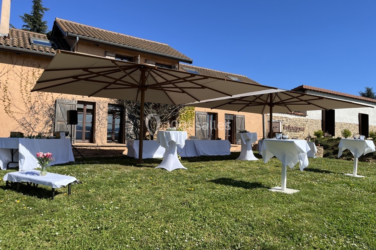 Tables hautes et parasols installés dans un jardin devant une maison en pierre sous un ciel dégagé.