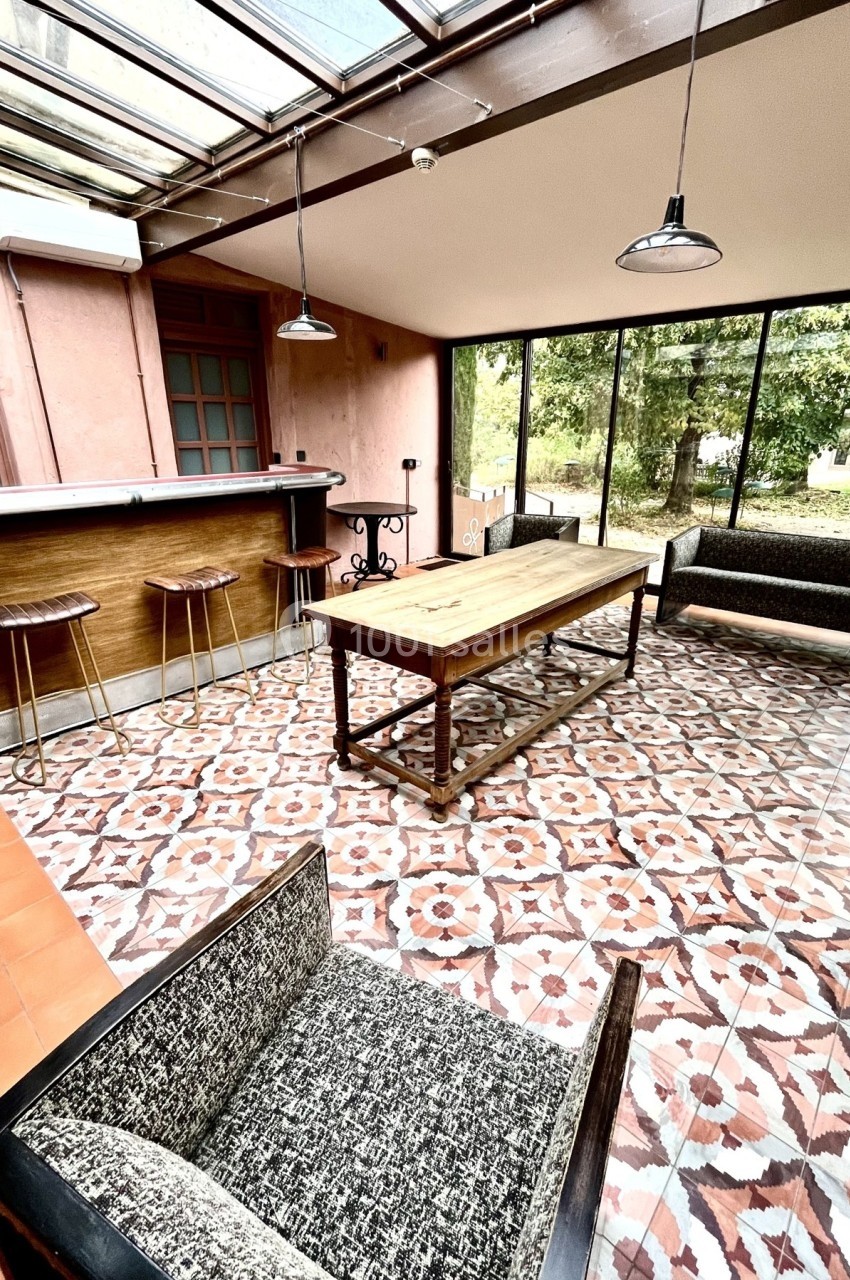 Salle lumineuse avec verrière, table en bois, chaises hautes près d'un bar, et canapés sur un sol à motifs géométriques.