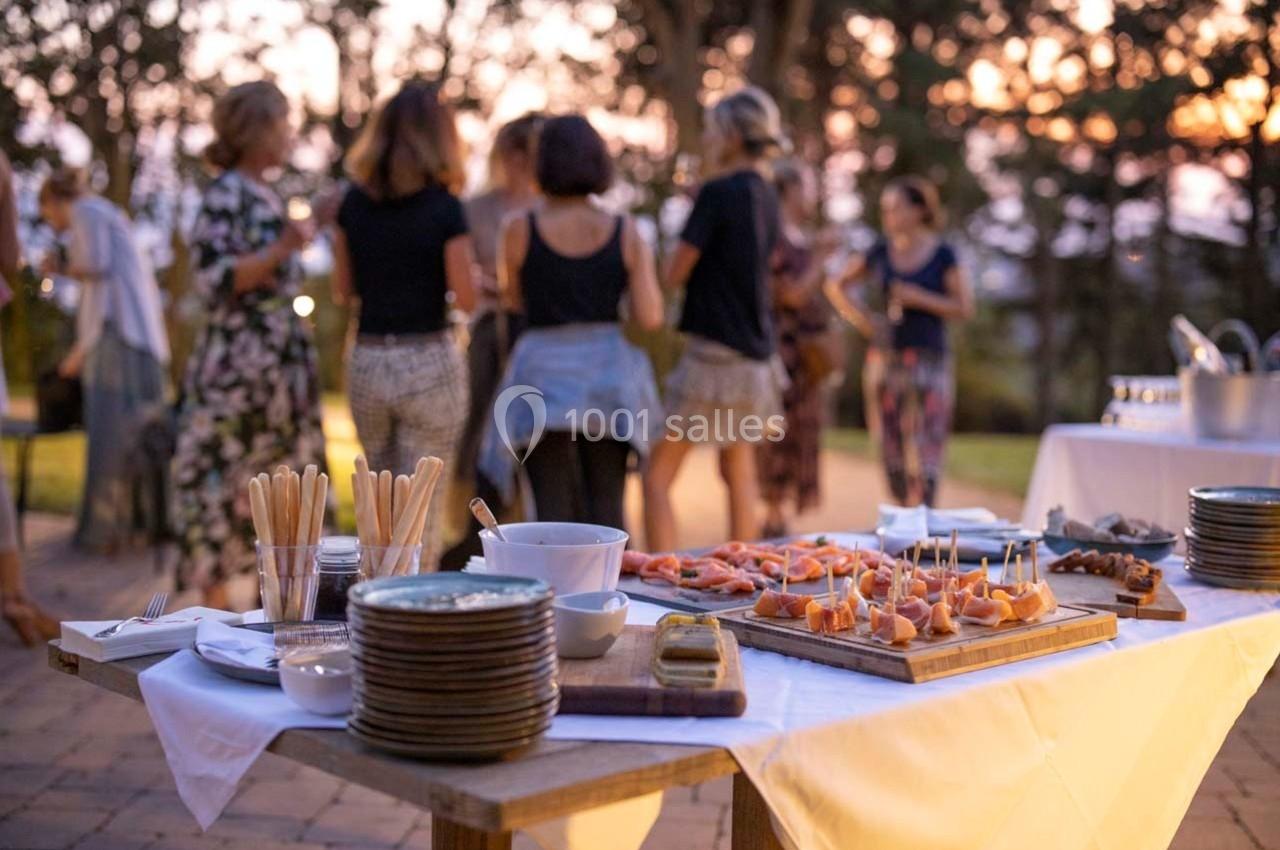 Table dressée avec des amuse-bouches et des assiettes, en arrière-plan des personnes discutent en extérieur au coucher du…