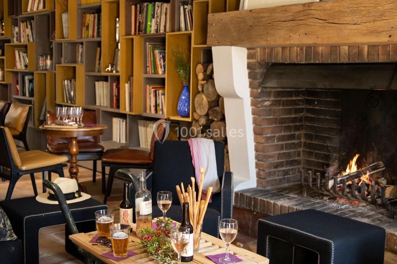 Salon chaleureux avec cheminée allumée, table basse garnie de boissons et snacks, entourée de fauteuils et étagères de…