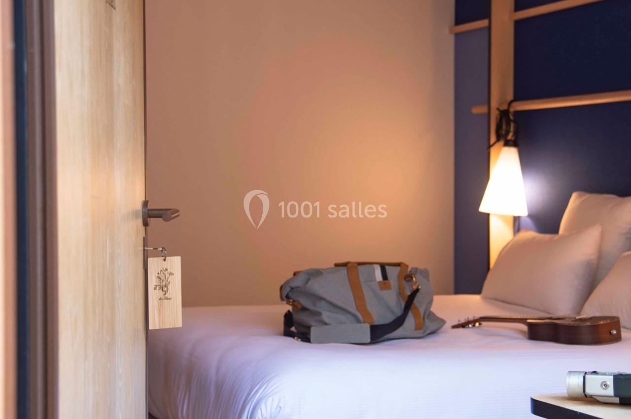 Chambre d'hôtel avec lit double, sac de voyage posé dessus, guitare et lampe allumée en arrière-plan.