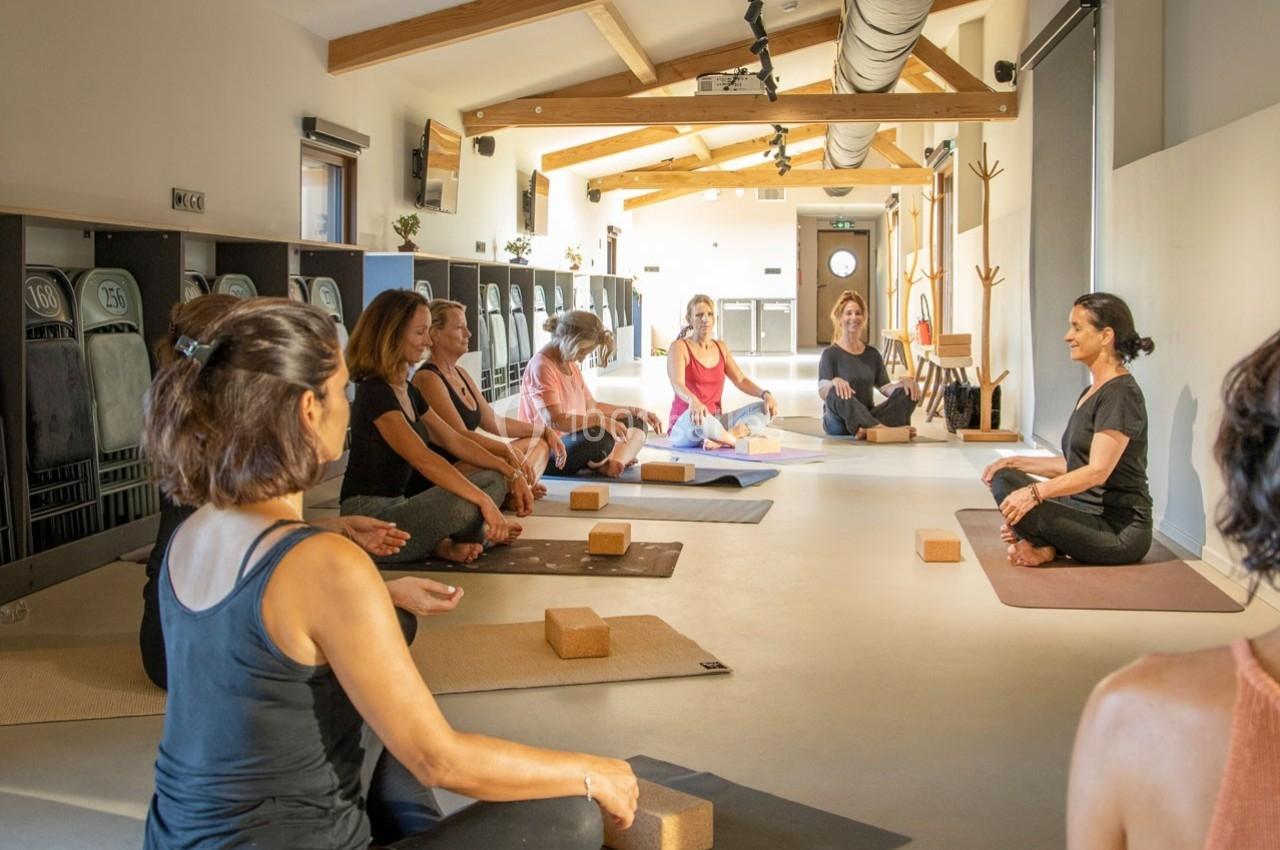 Un groupe de personnes assises sur des tapis de yoga dans une salle lumineuse, écoutant une instructrice.
