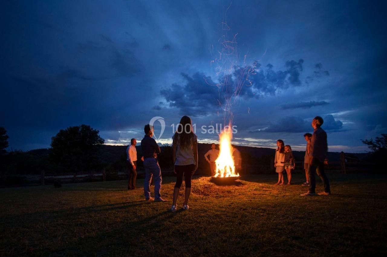 Des personnes debout autour d'un feu de camp dans un champ au crépuscule, sous un ciel nuageux.