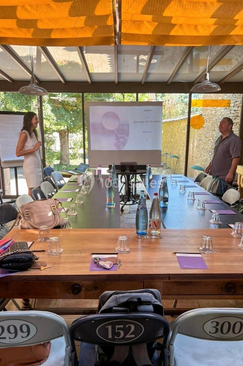 Salle de réunion lumineuse avec une grande table en bois, des chaises numérotées et un écran de présentation allumé.