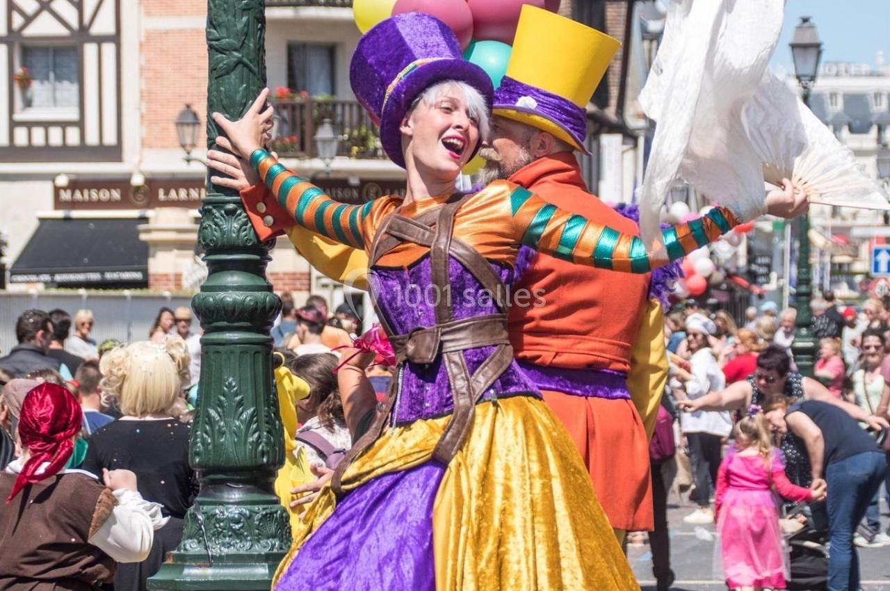 Femme en costume coloré et chapeau violet dans une parade festive, tenant un lampadaire près de ballons multicolores.