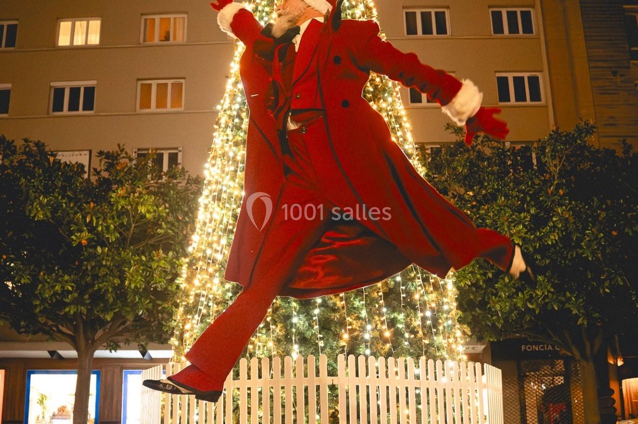 Un homme déguisé en Père Noël saute devant un grand sapin de Noël illuminé, entouré d'une clôture blanche.