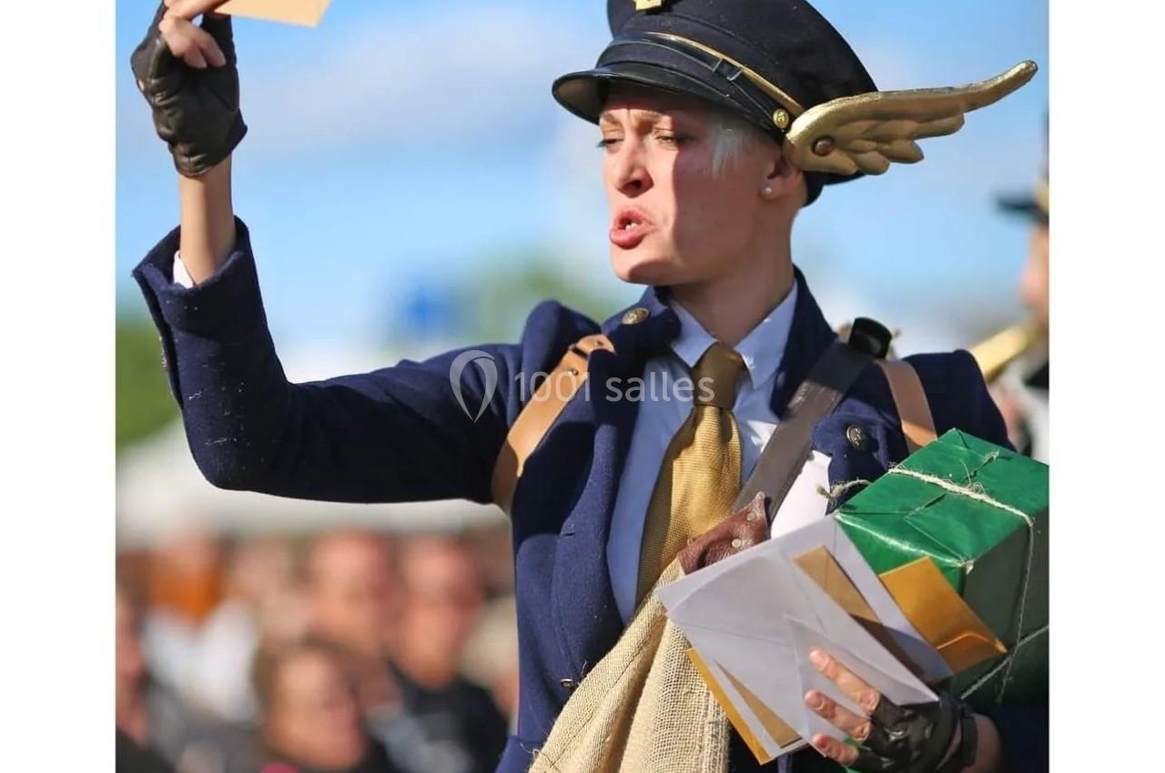 Une personne déguisée en facteur d'époque, tenant une lettre en l'air, avec un sac rempli de courrier.