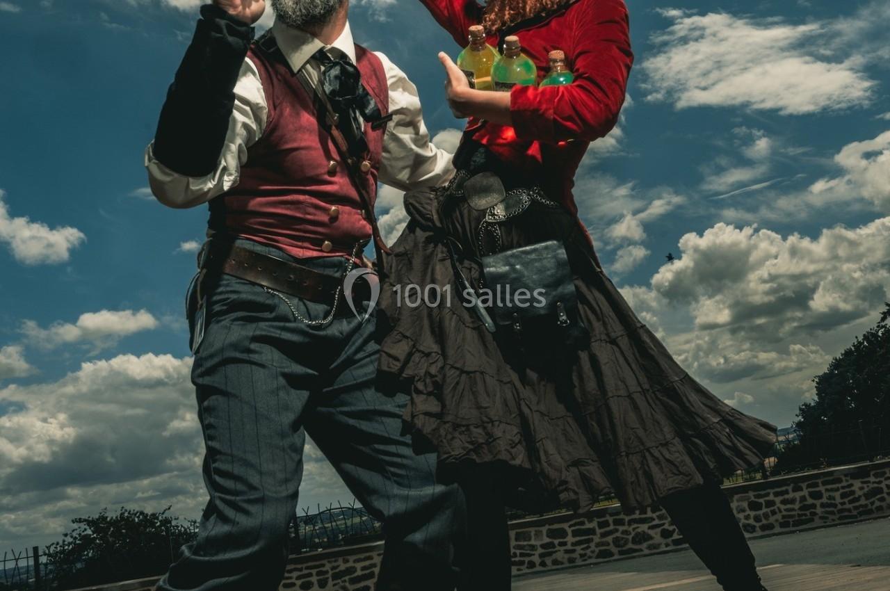 Deux personnes en costumes steampunk posent en plein air sous un ciel nuageux, l'une tenant une corne, l'autre une bouteille.