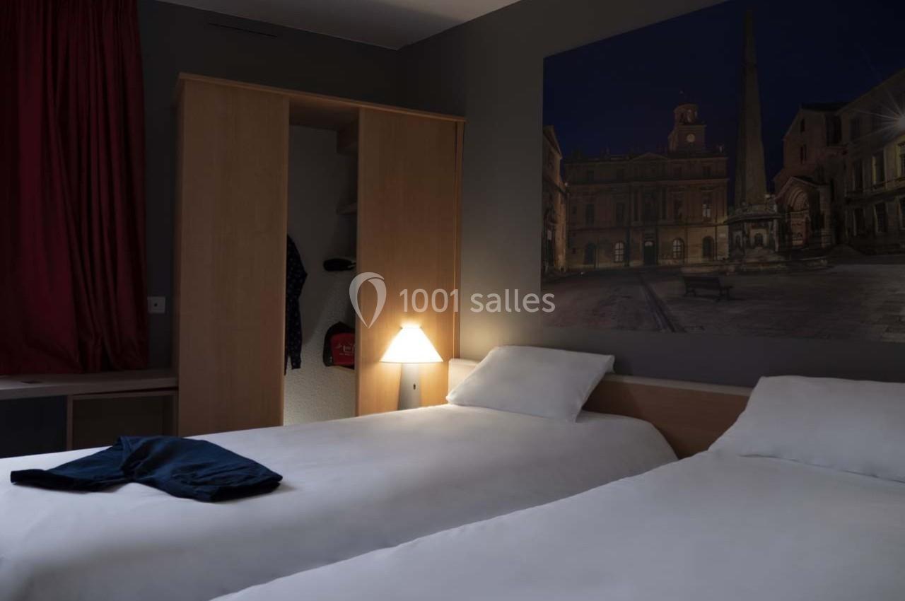 Deux lits simples dans une chambre d'hôtel sombre, éclairée par une lampe de chevet, avec un tableau mural en arrière-plan.