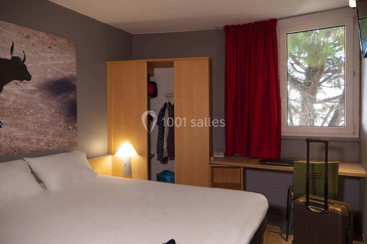 Chambre d'hôtel avec lit double, armoire ouverte, bureau, valise au sol, et fenêtre avec rideaux rouges.