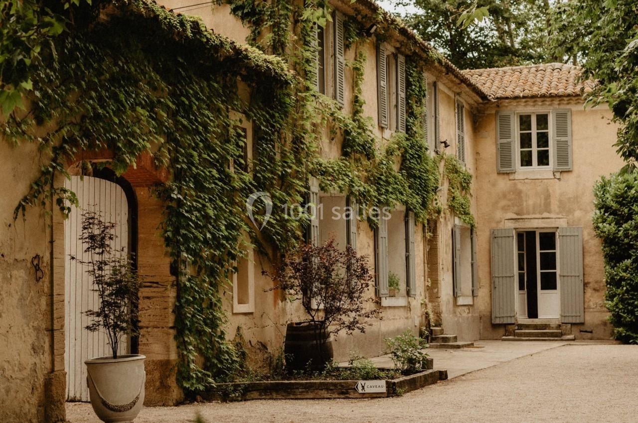 Façade d'une maison ancienne en pierre, couverte de lierre, entourée d'arbres et d'un jardin avec des pots de plantes.