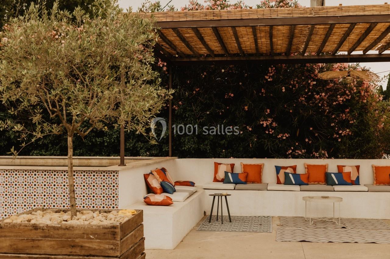 Terrasse extérieure avec banquettes blanches, coussins colorés, olivier en pot et pergola en bois.