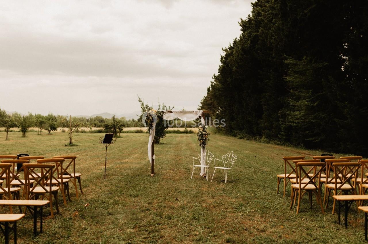 Chaises et arche décorée pour une cérémonie en plein air, disposées dans un champ bordé d'arbres.