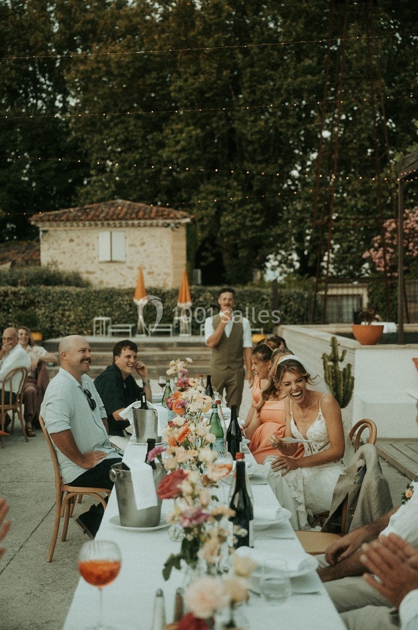 Des convives souriants assis à une table décorée de fleurs lors d'un repas en extérieur, sous un ciel dégagé.