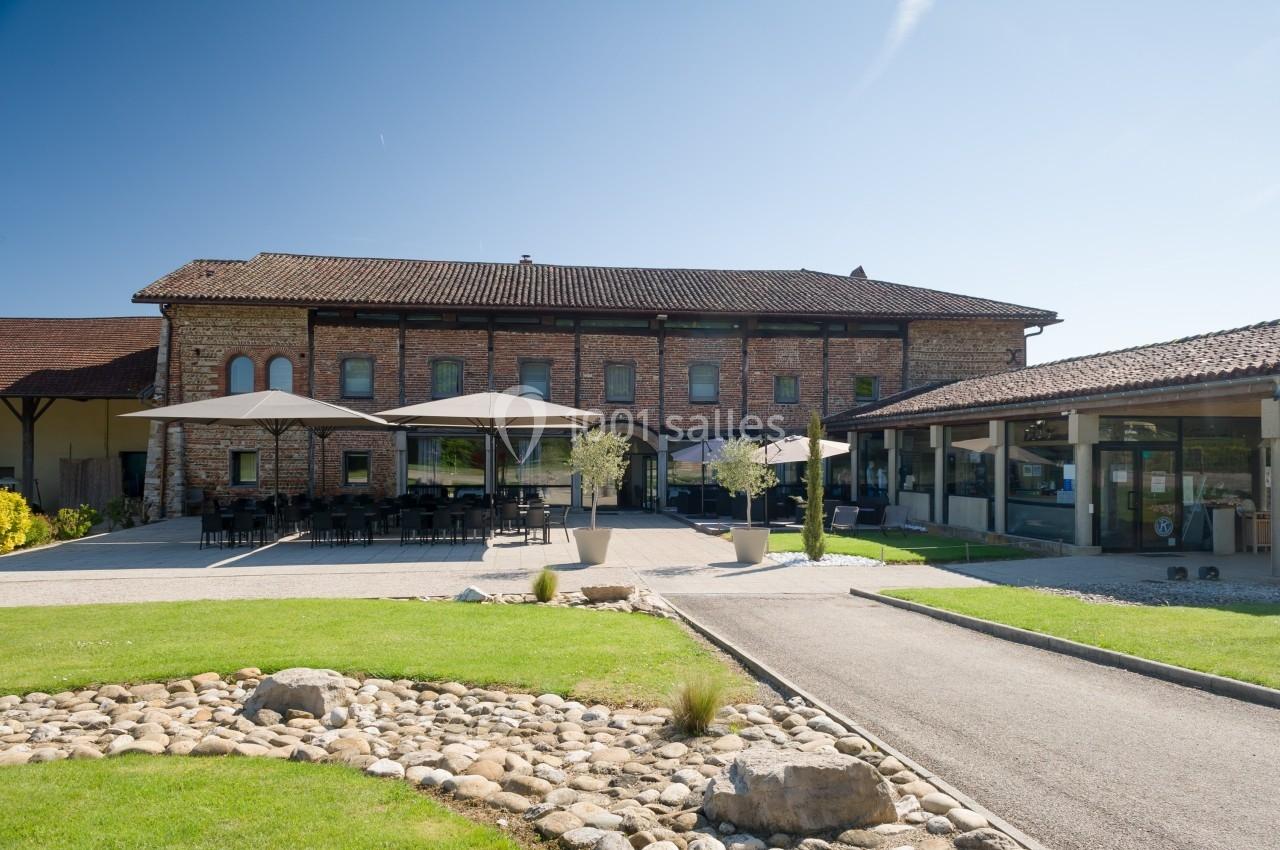 Bâtiment en briques avec terrasse ombragée, tables et chaises, entouré d'un jardin avec allée et pelouse.
