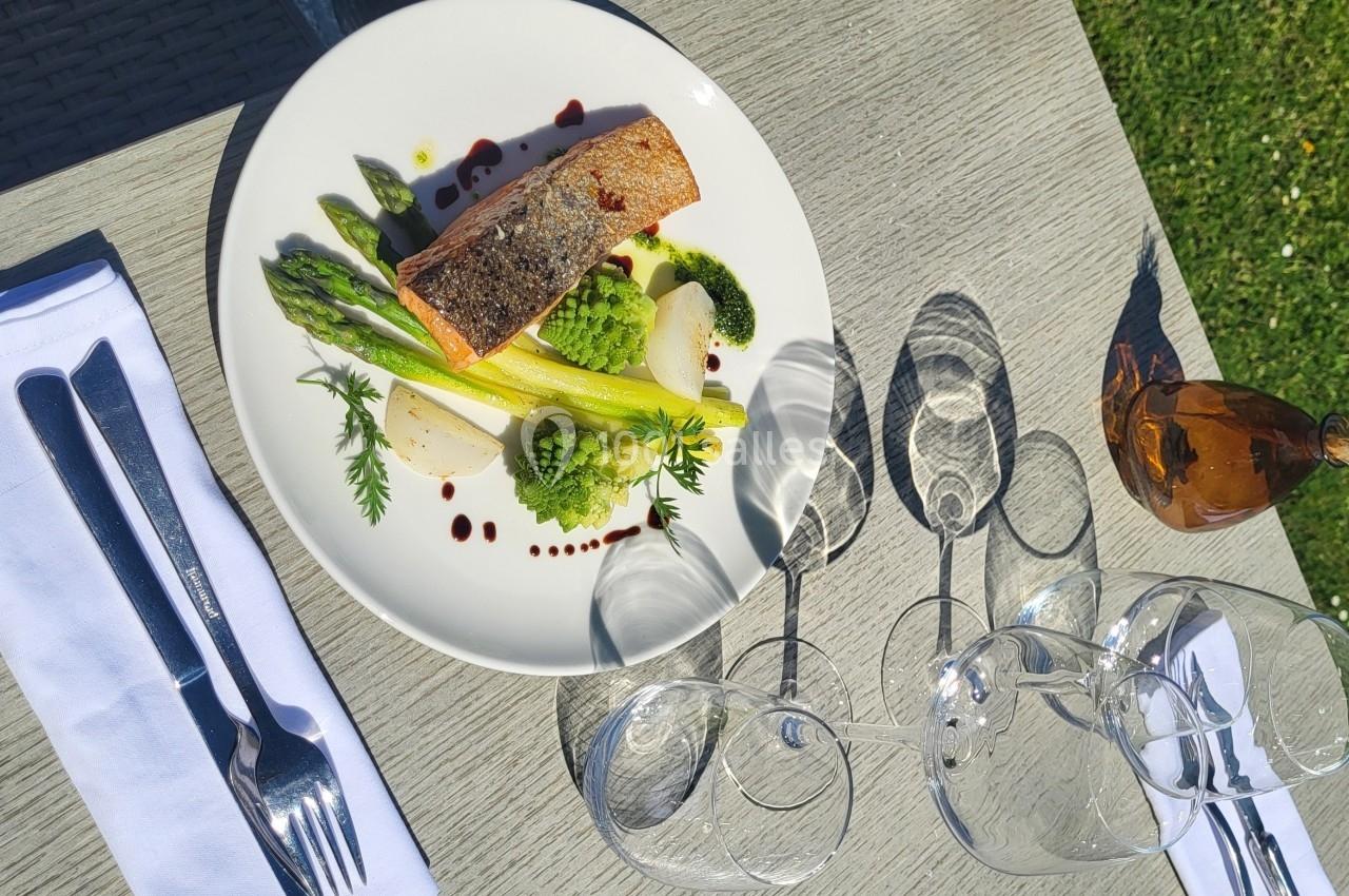 Assiette de saumon grillé avec asperges et sauce, servie sur une table extérieure avec couverts et verres.