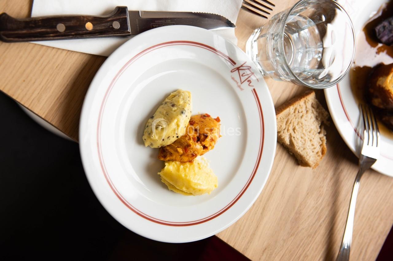 Assiette contenant trois quenelles de purées colorées, posée sur une table avec un couteau, un verre d'eau et du pain.