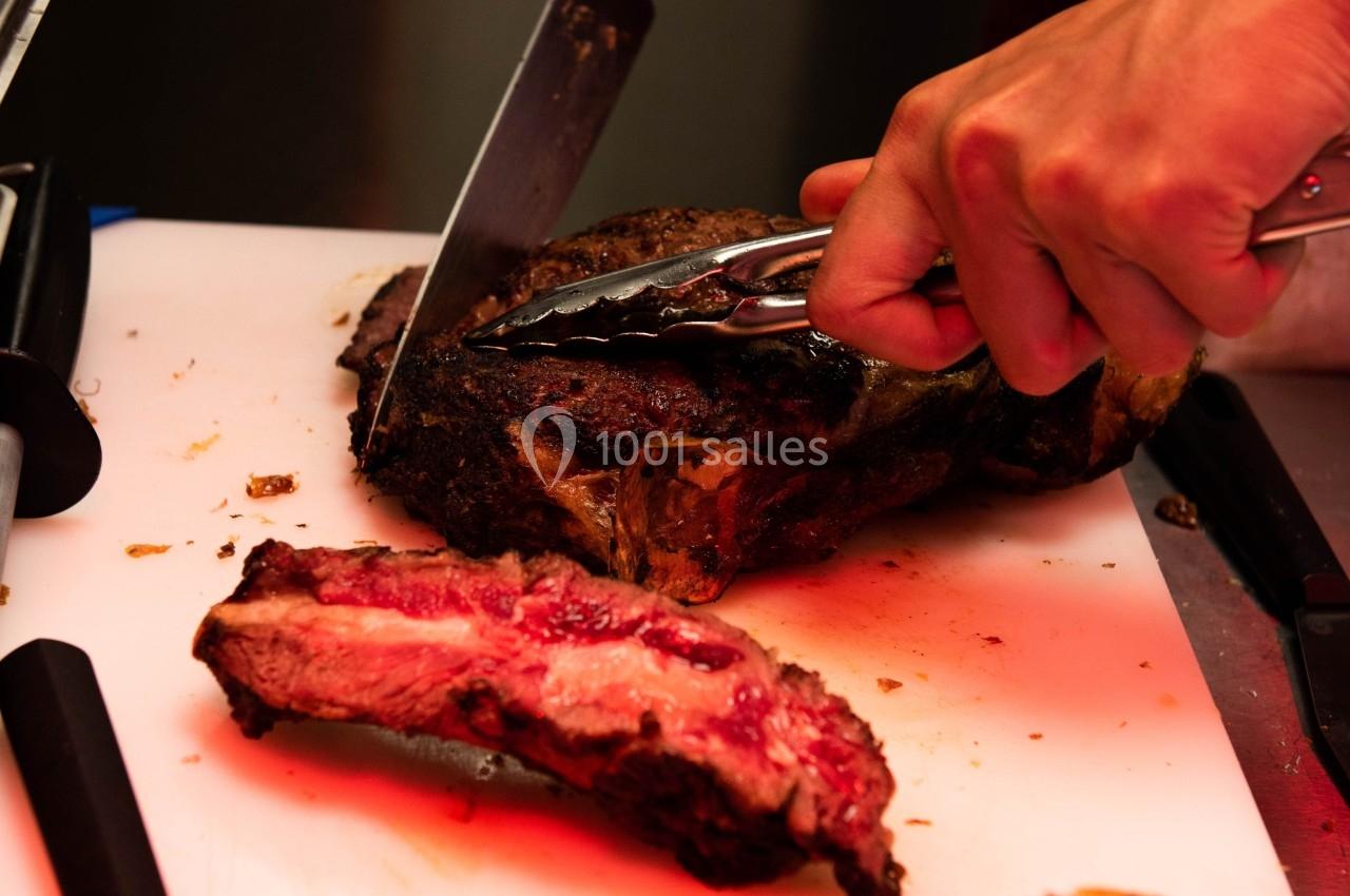 Une main découpe un morceau de viande grillée sur une planche blanche à l'aide d'un couteau et d'une pince.