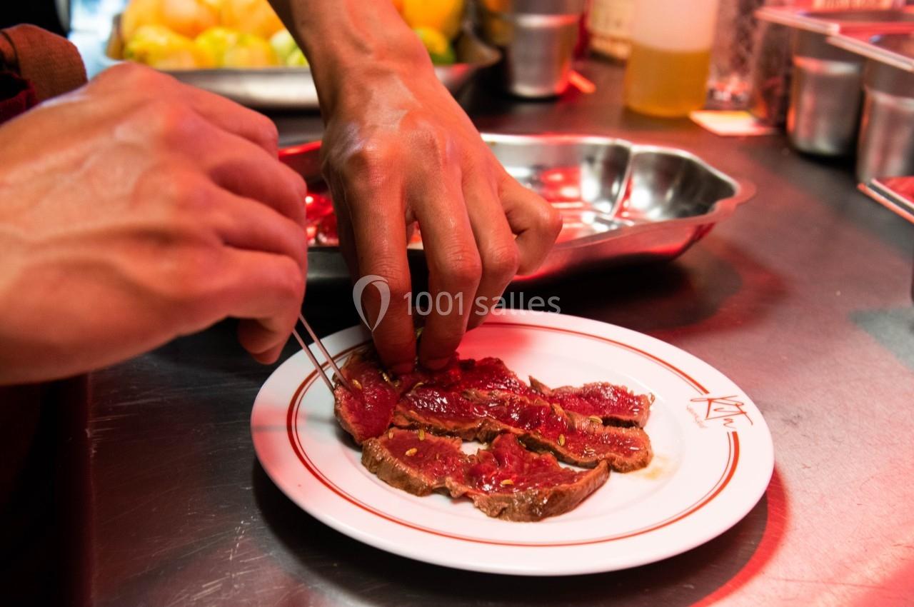 Mains dressant des tranches de viande rouge sur une assiette blanche dans une cuisine professionnelle.
