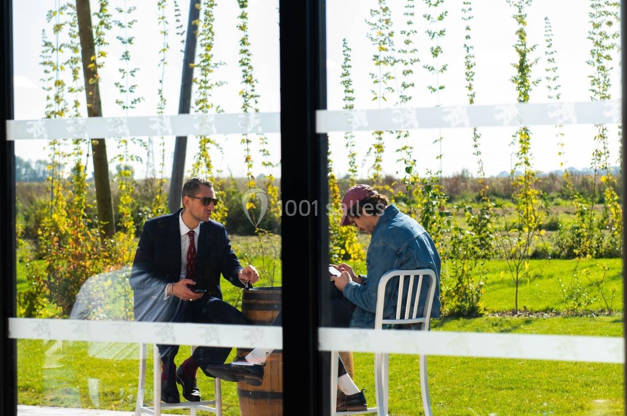 Deux hommes discutent à une table en extérieur, près d'une grande baie vitrée, avec un paysage verdoyant en arrière-plan.