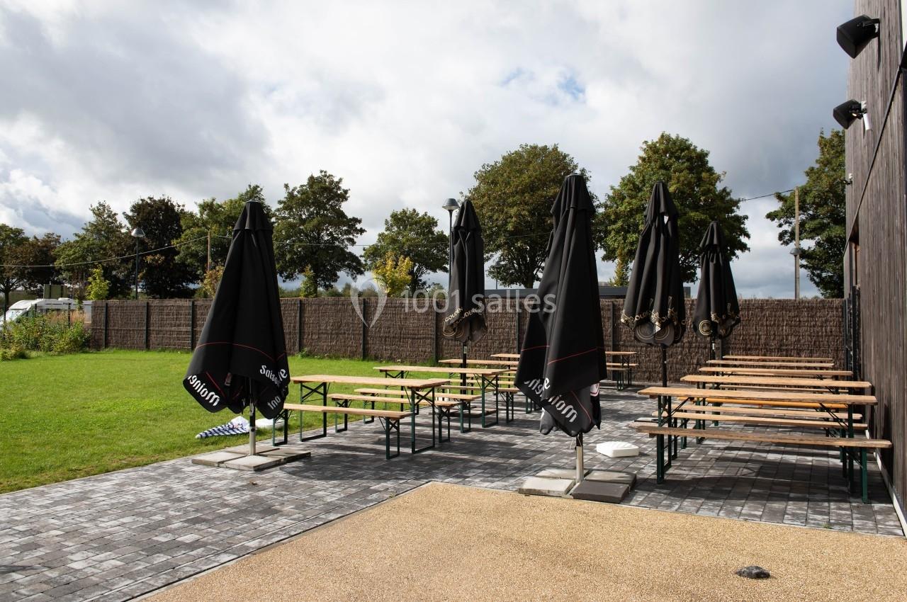 Terrasse extérieure avec tables en bois et parasols noirs, entourée de pelouse et d'arbres sous un ciel nuageux.