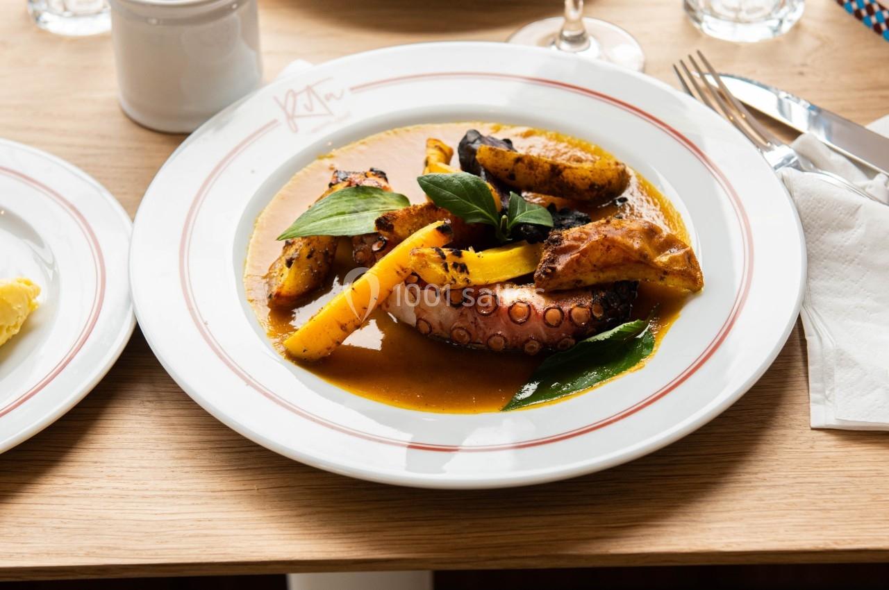 Assiette de poulpe grillé avec légumes et sauce, servie sur une table en bois avec un verre de vin rouge.