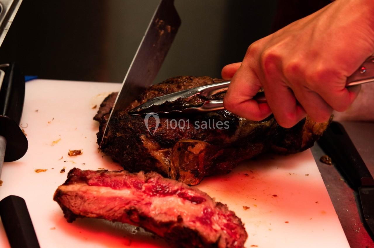 Un cuisinier découpe un morceau de viande grillée sur une planche blanche avec un couteau et une pince.