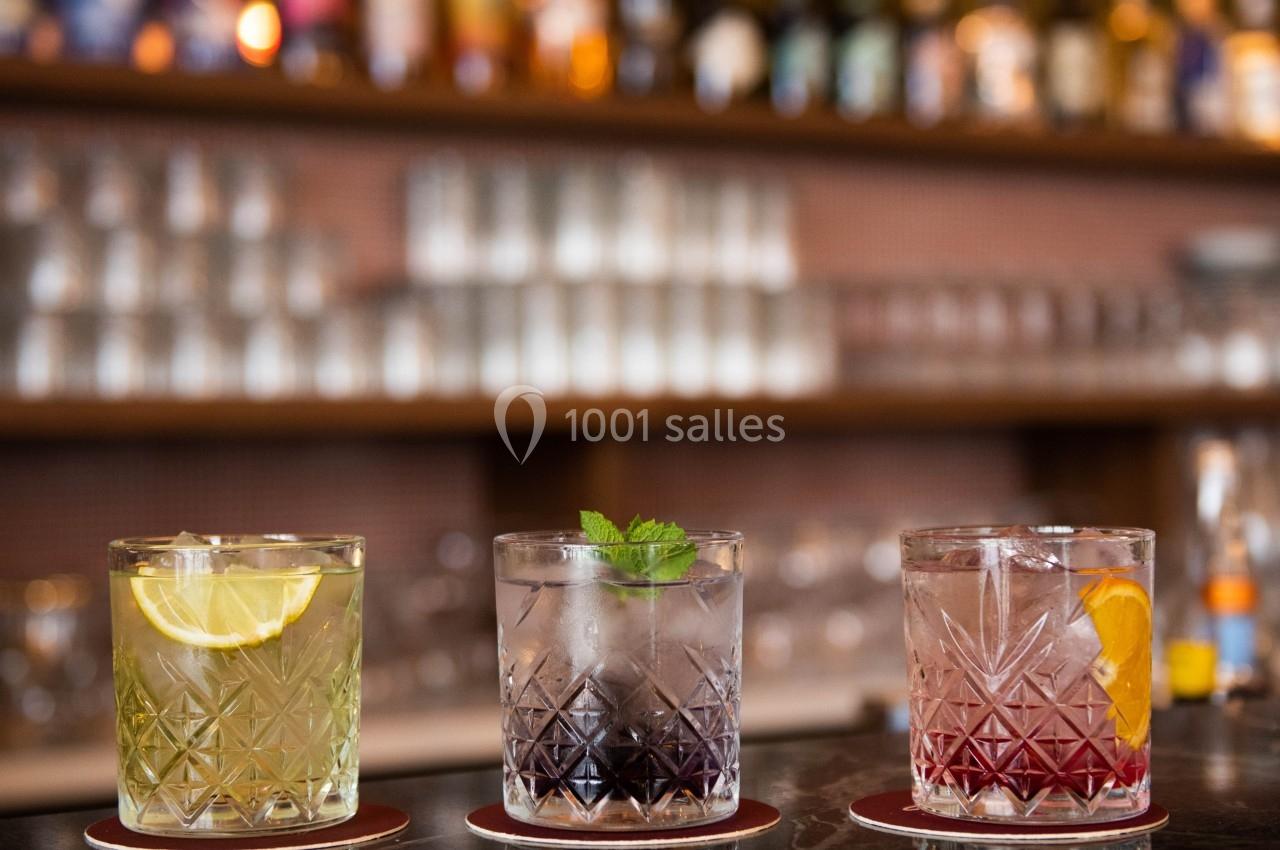 Trois cocktails colorés servis dans des verres en cristal, posés sur un comptoir devant un bar en arrière-plan flou.