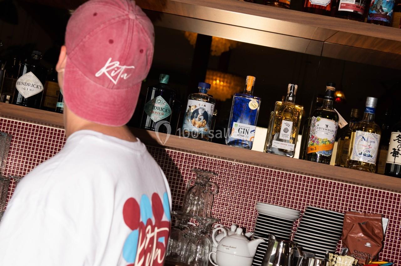Un homme de dos portant une casquette et un t-shirt devant un bar avec des bouteilles d'alcool alignées sur des étagères.