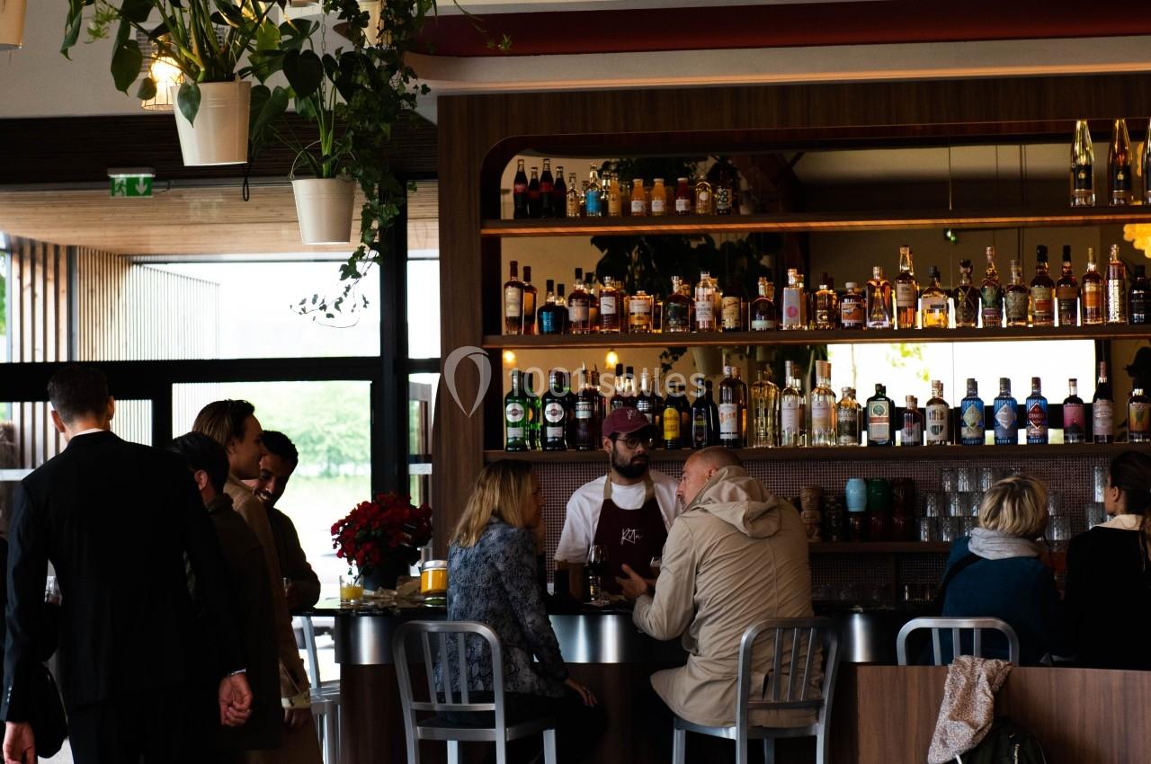Intérieur d'un bar-restaurant avec des clients au comptoir, des tables dressées et des plantes suspendues.