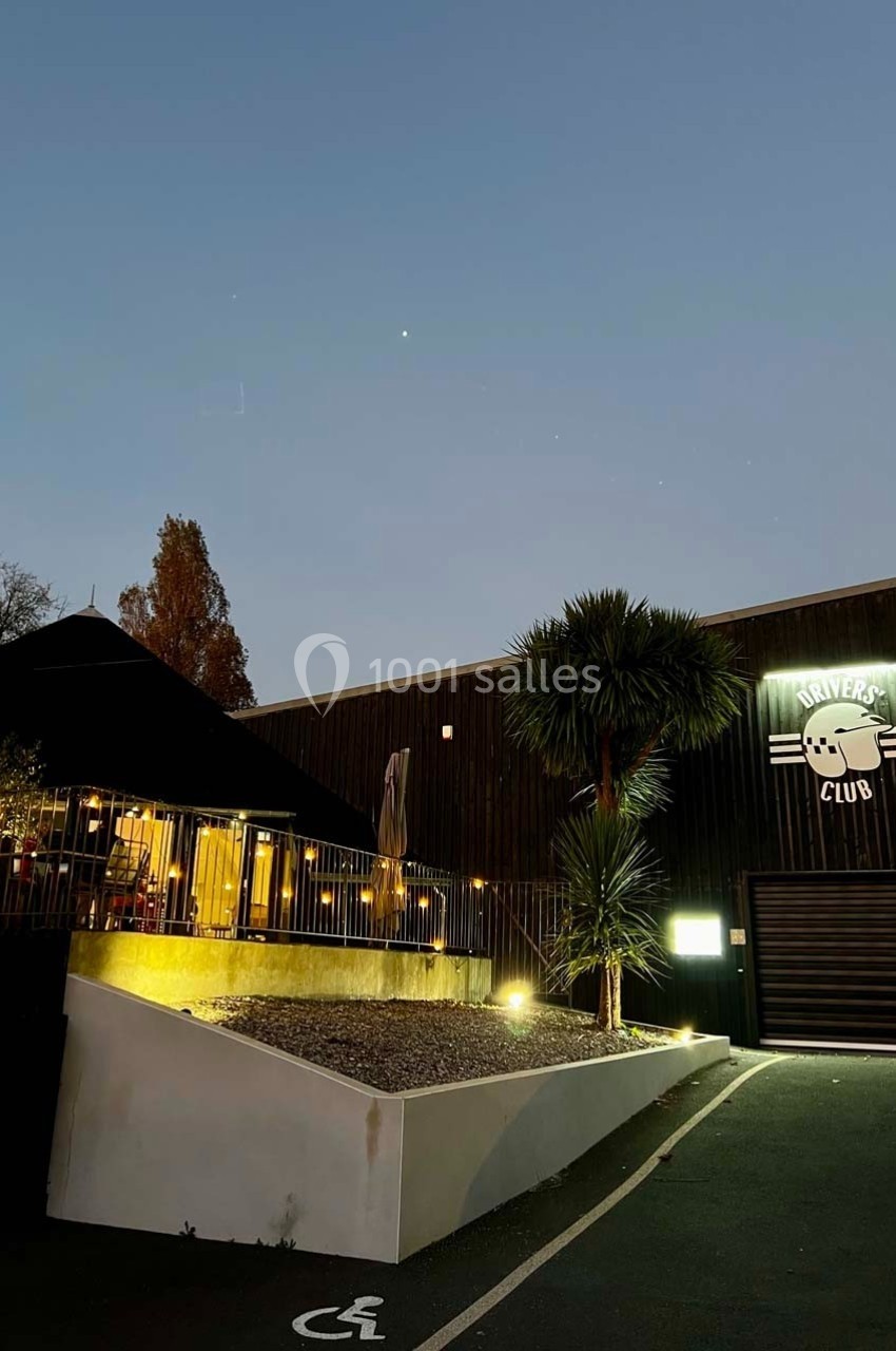 Façade d'un bâtiment en soirée avec enseigne lumineuse, terrasse éclairée et emplacement de parking accessible.