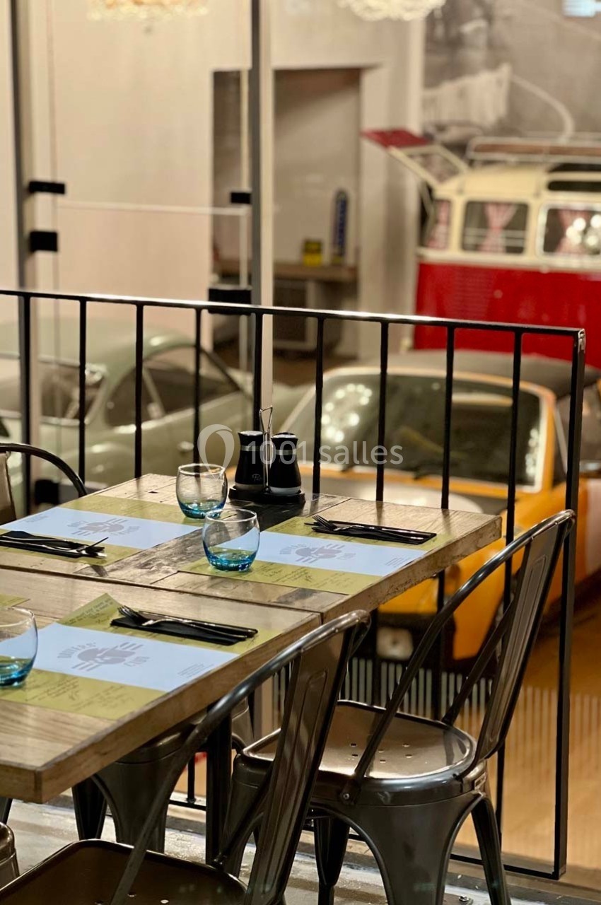 Table en bois dressée avec couverts et verres, située dans un restaurant avec vue sur des voitures anciennes en arrière-plan.