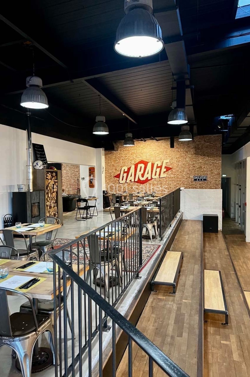 Salle de restaurant au style industriel avec murs en briques, tables en métal et bois, et enseigne ’Garage’ au fond.