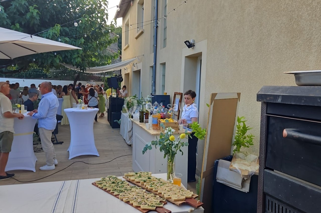 Terrasse extérieure avec buffet, tables hautes et invités discutant près d'un bar sous des guirlandes lumineuses.