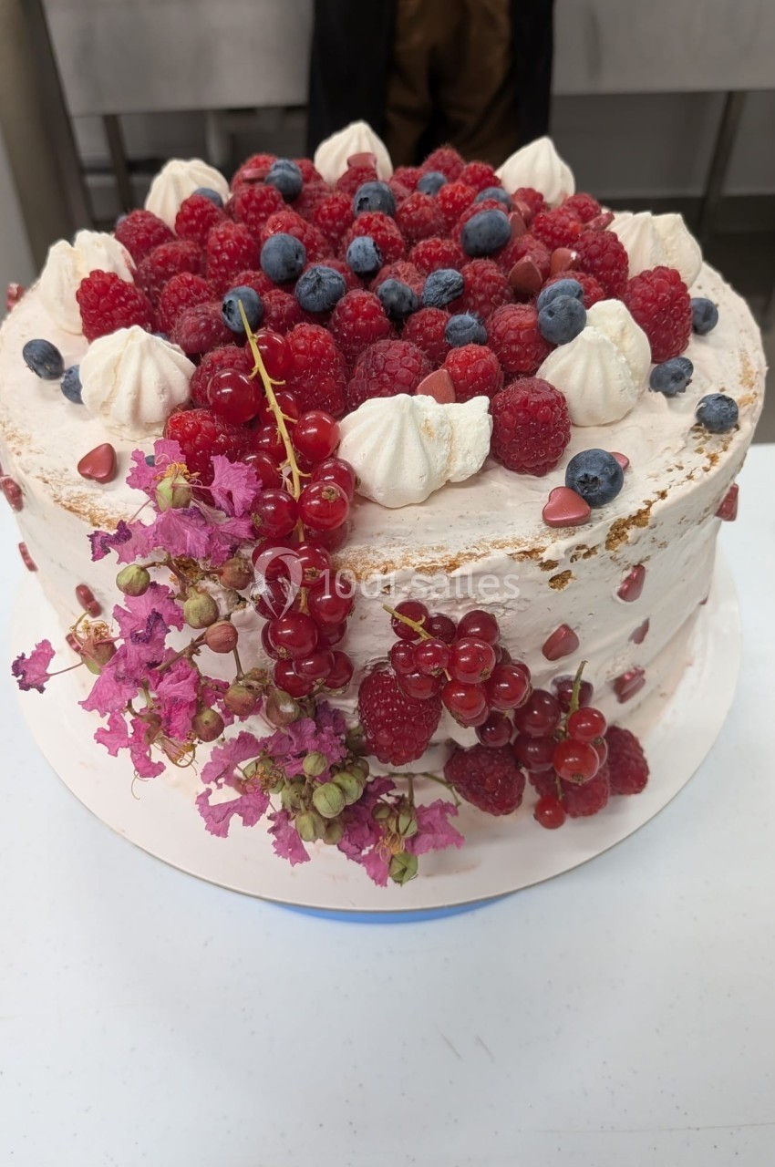 Gâteau décoré de fruits rouges, myrtilles, groseilles, meringues et fleurs roses sur une table blanche.