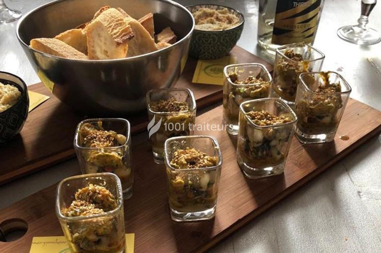 Plateau en bois avec verrines garnies, bol de pain tranché et bouteille de vin mousseux en arrière-plan.