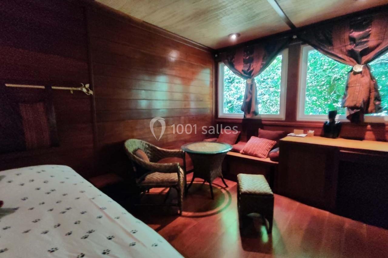 Chambre en bois avec lit, fauteuils en osier, table ronde, bureau et fenêtres donnant sur un paysage verdoyant.